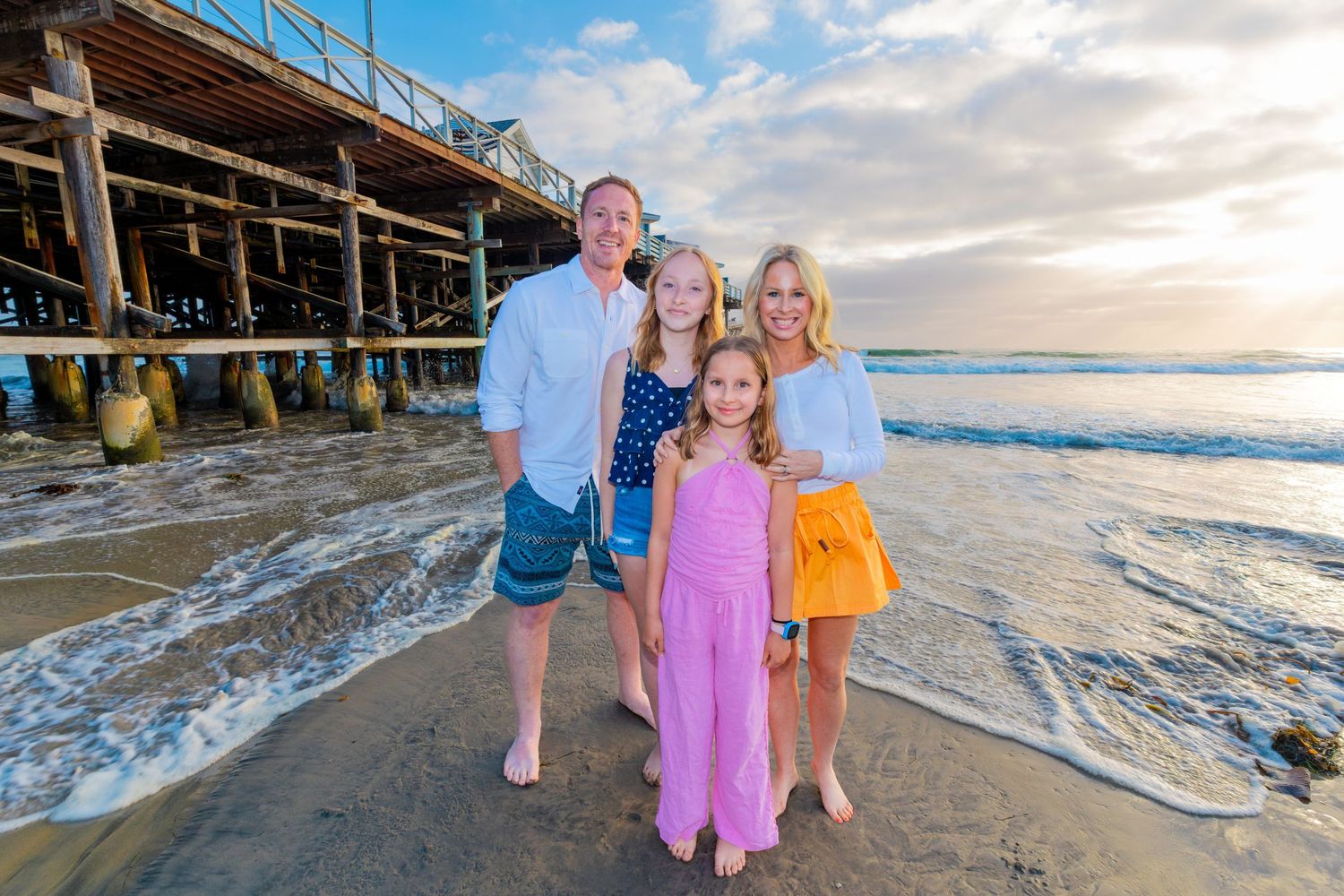 San Diego family portrait photography of a family of four standing on the beach next to a wooden pier with ocean waves.