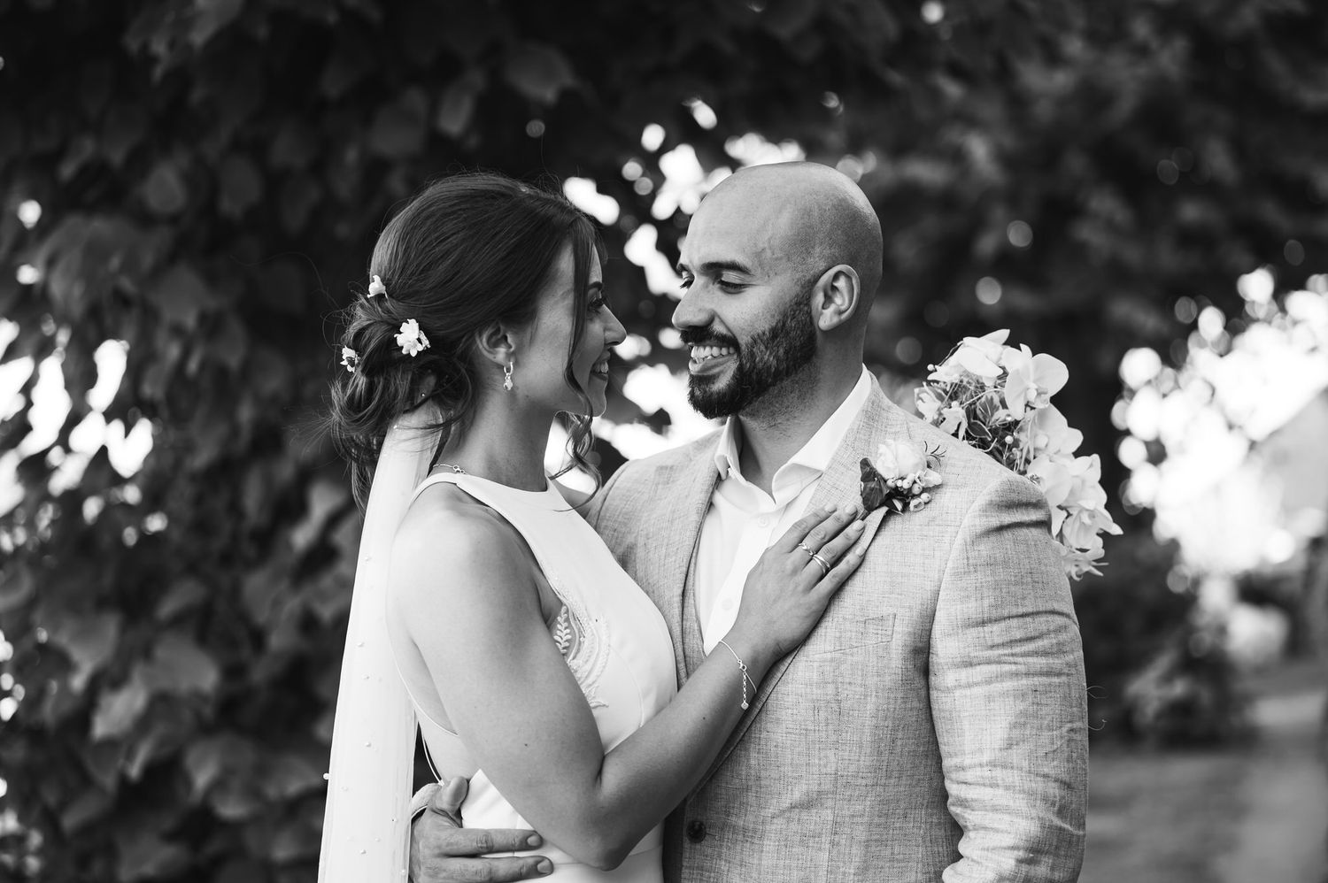 Black and white photo of a smiling couple in wedding attire, groom in suit with boutonniere, bride holding his lapel.