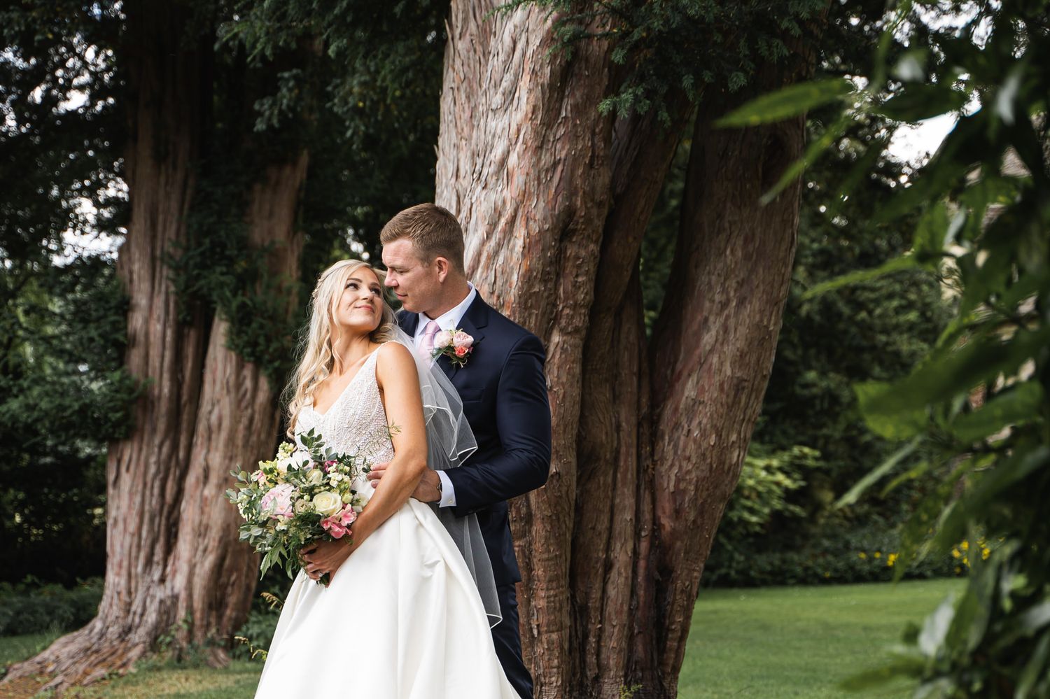 Bride and groom embracing by ancient trees in a lush garden, bride holding a pastel bouquet in a white gown.