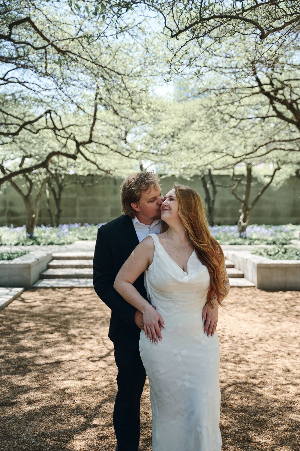 Bride and groom embrace under blooming trees in an outdoor courtyard setting on their wedding day.