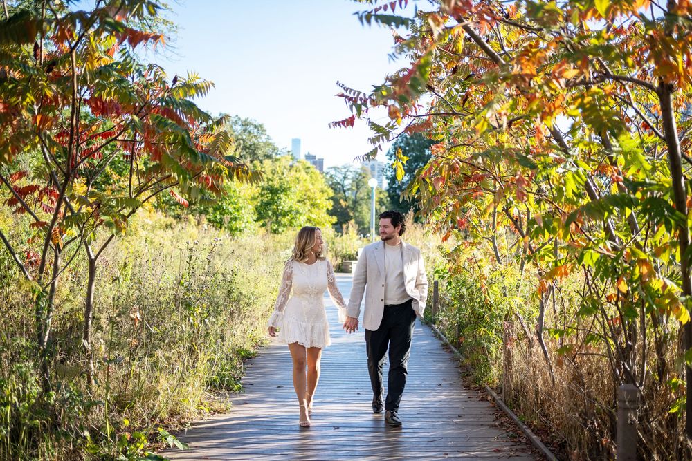 Couple walks hand-in-hand on a wooden boardwalk surrounded by vibrant autumn foliage with city skyline in background.