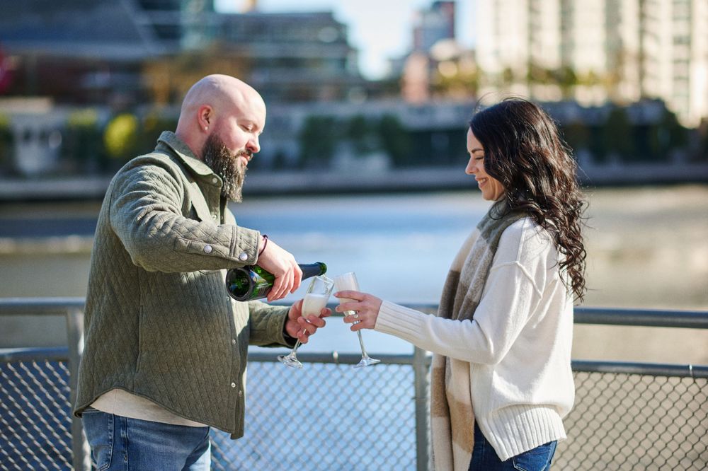 Man pouring champagne into a woman's glass on a waterfront bridge during a romantic outdoor celebration.