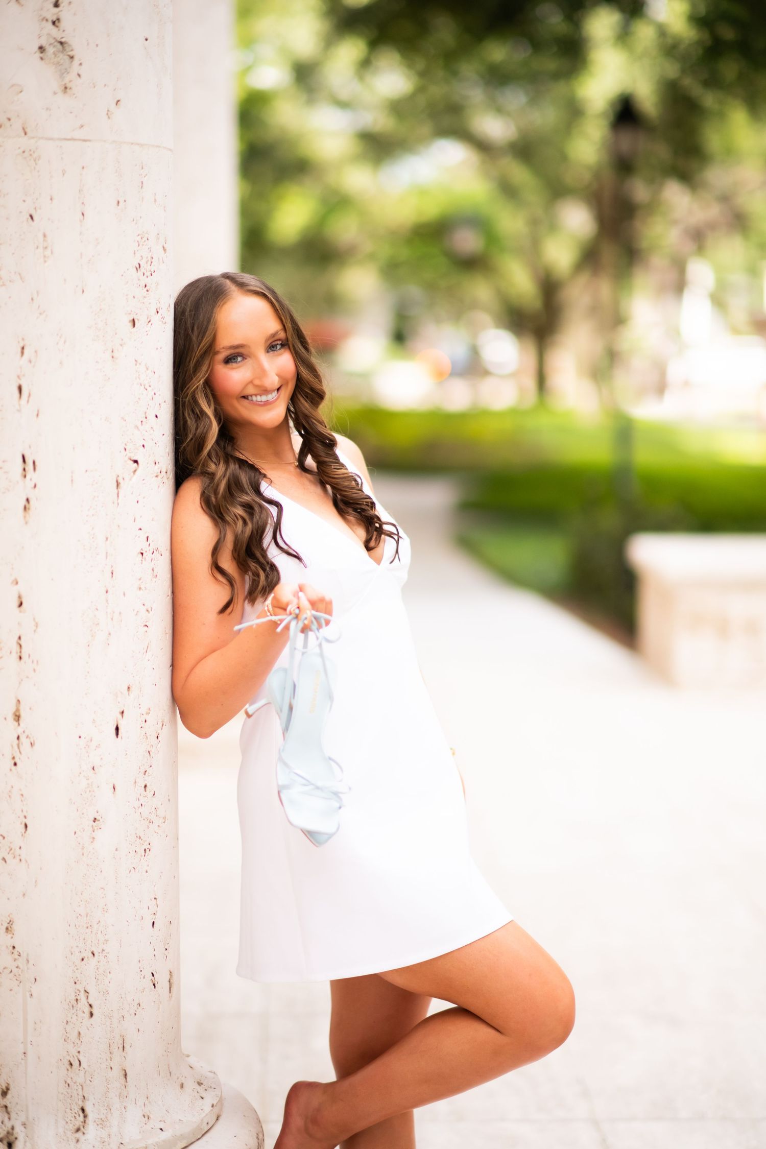 Young woman in white dress leaning against a marble column, smiling outdoors with green trees in background.