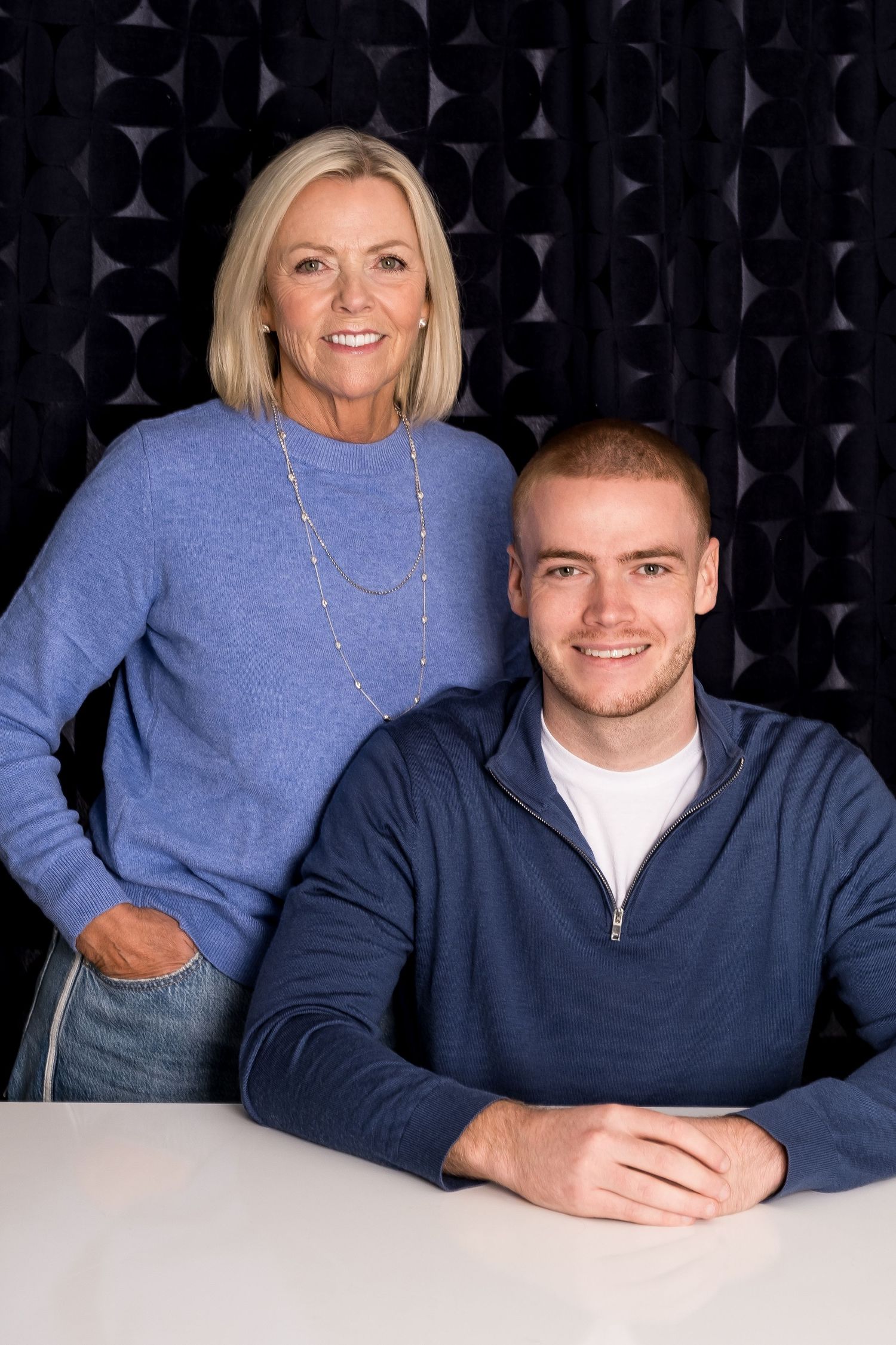 A smiling blonde woman in a blue sweater stands behind a young man in a navy zip-up at a white table.