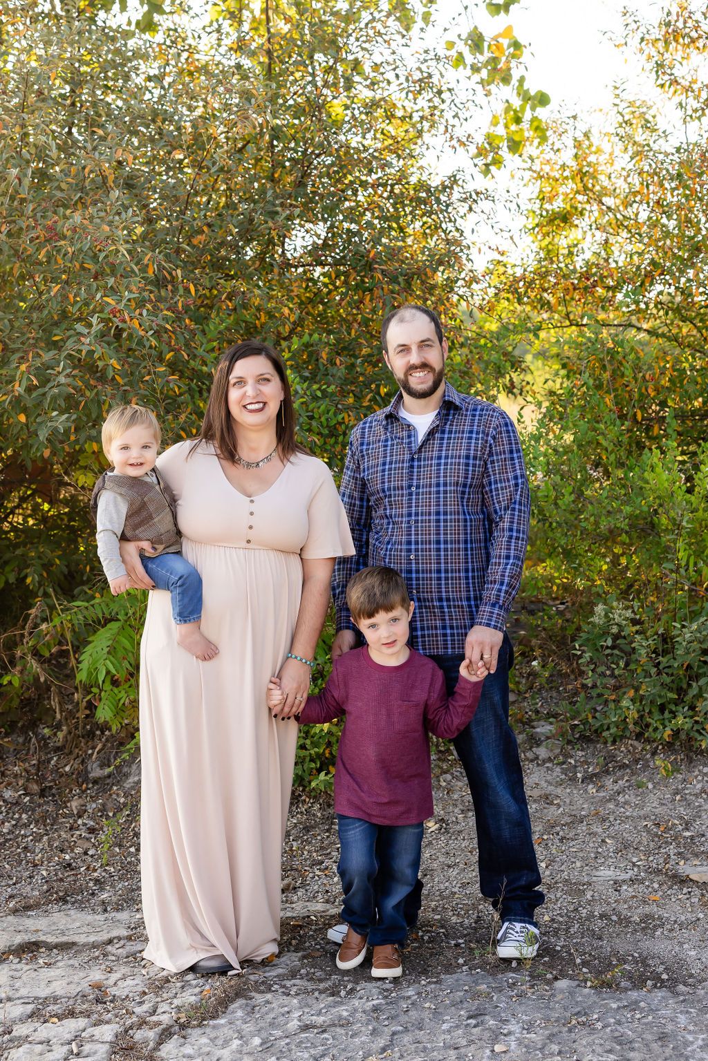 Family of four posing outdoors among green foliage, mother holding toddler in a blush maxi dress, father and son holding hands.