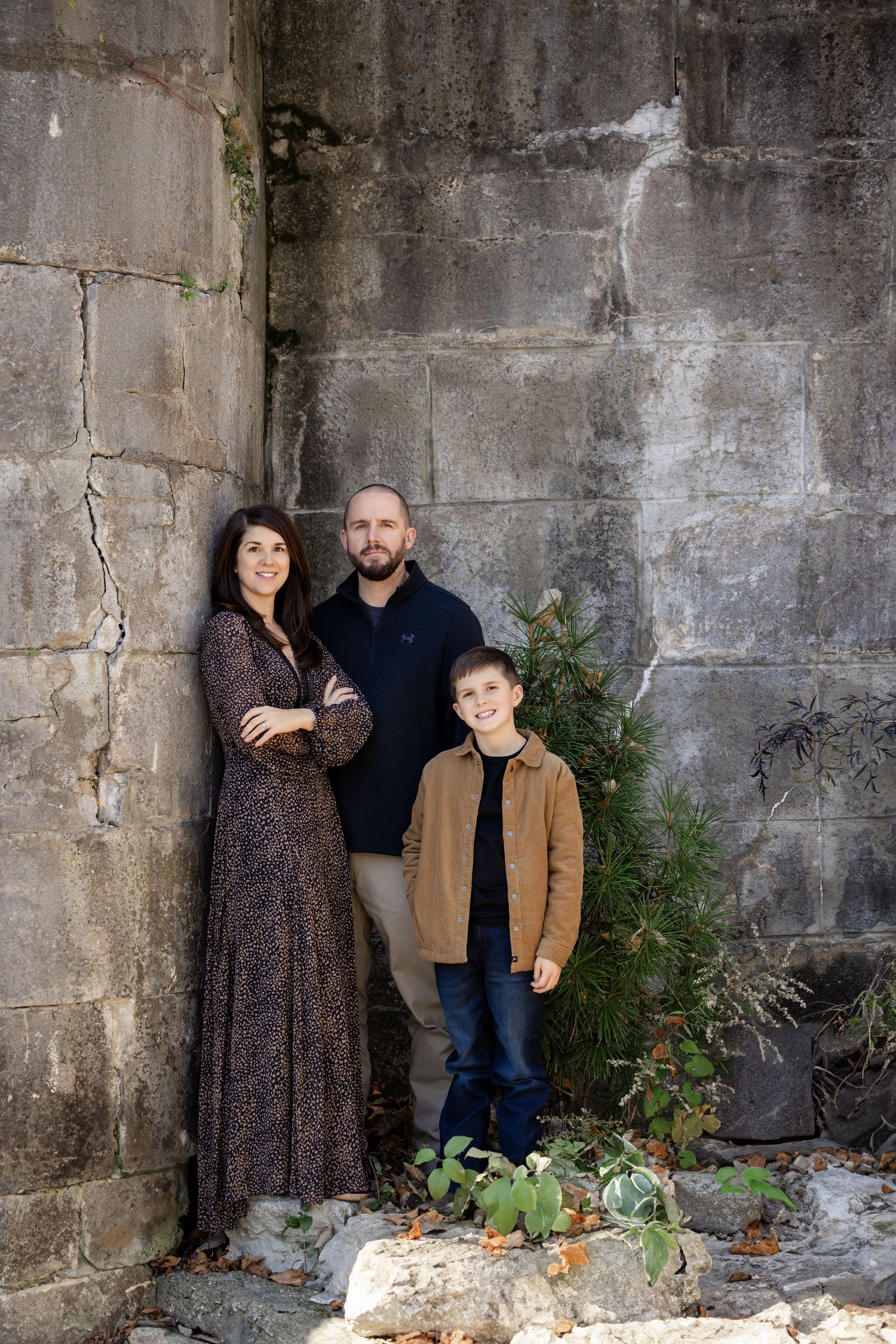 Family of three posing against a stone wall outdoors, parents and young boy smiling in coordinated fall outfits.