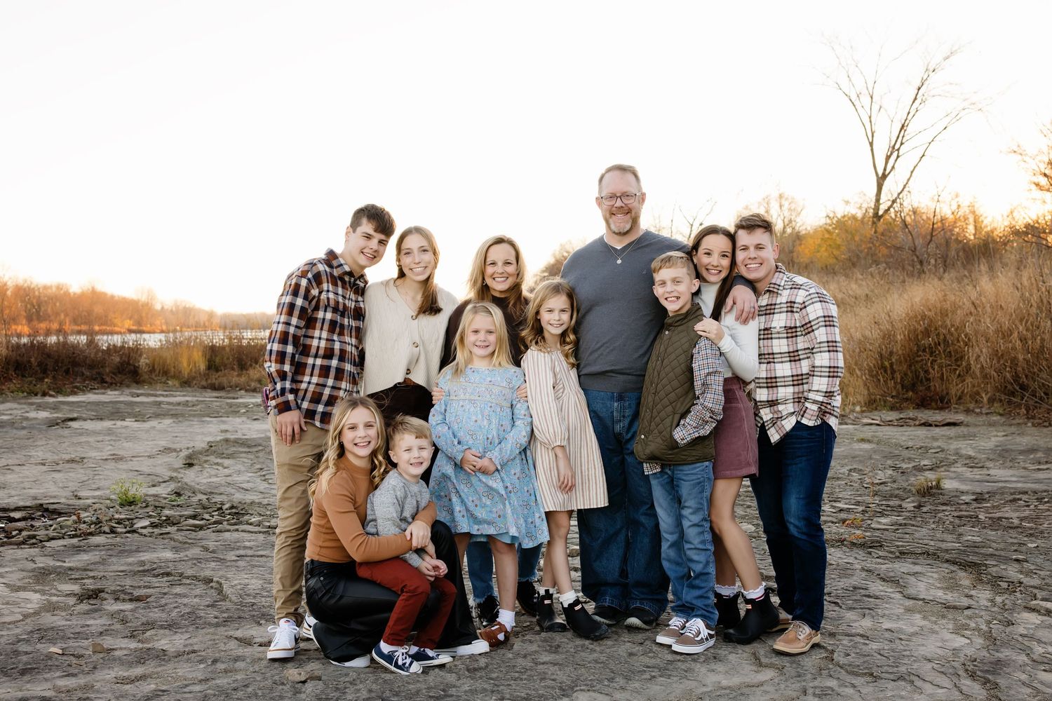 Large family group portrait outdoors during golden hour, posing together on rocky terrain with autumn trees in background.