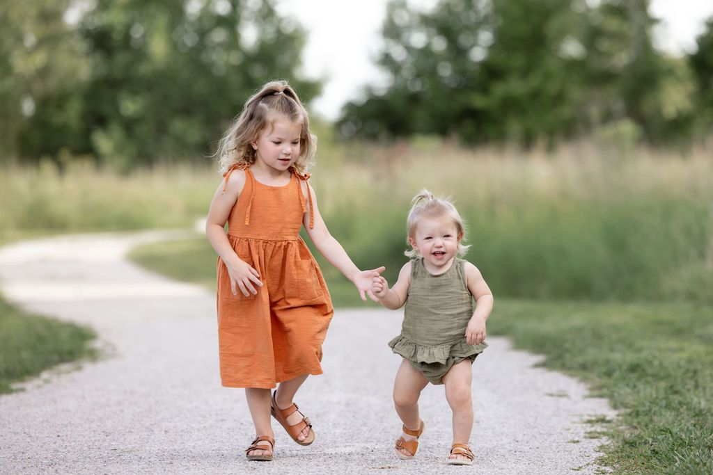 Two young sisters holding hands and walking together on a park path in matching outfits.