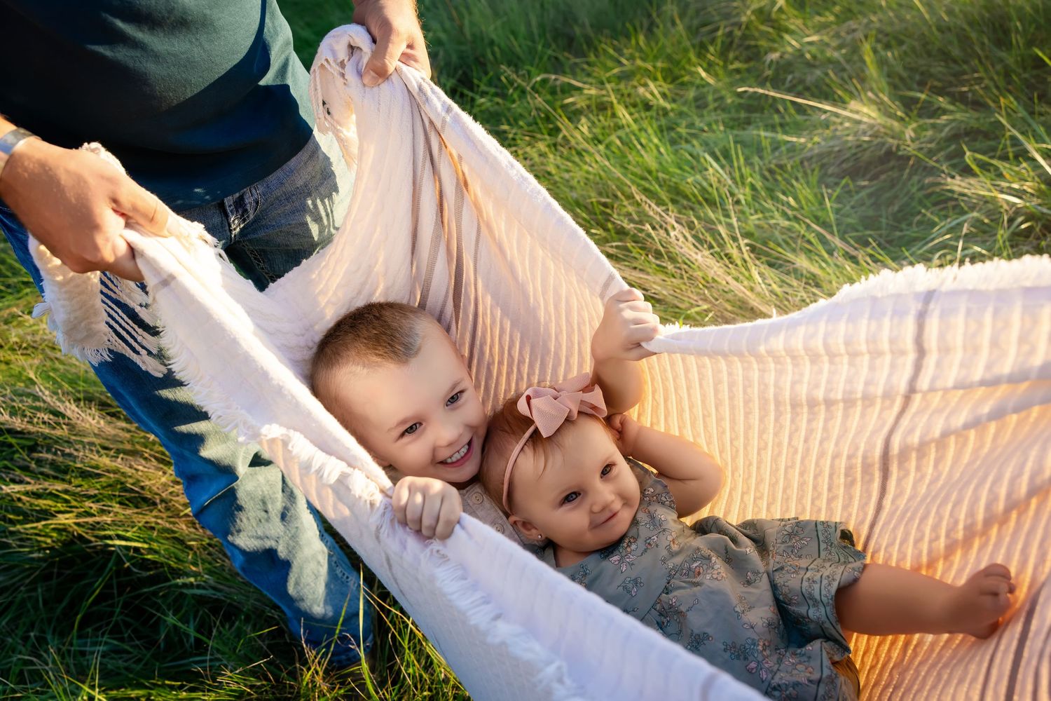 Two babies lying in a blanket hammock held by adults in a sunny green grass field.