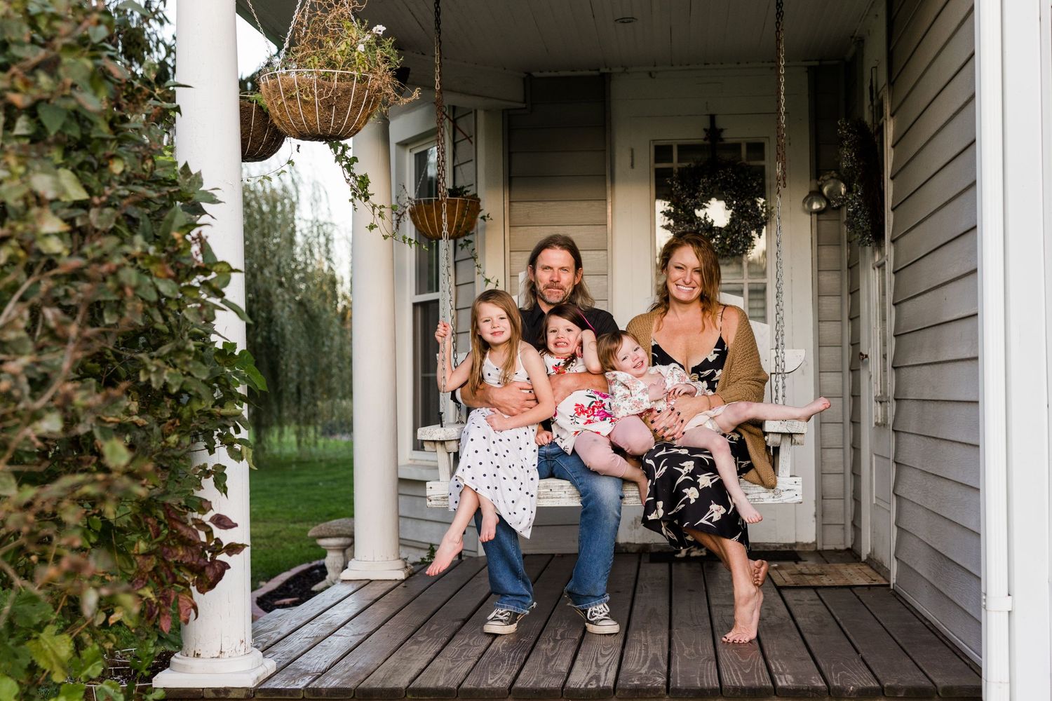 Family of five poses on a charming farmhouse porch, parents with three young daughters smiling warmly on a cloudy day.