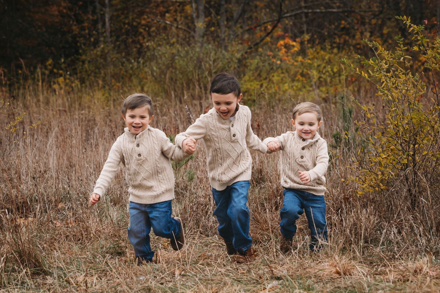 Three young children in matching cream sweaters and jeans run through an autumn field holding hands.