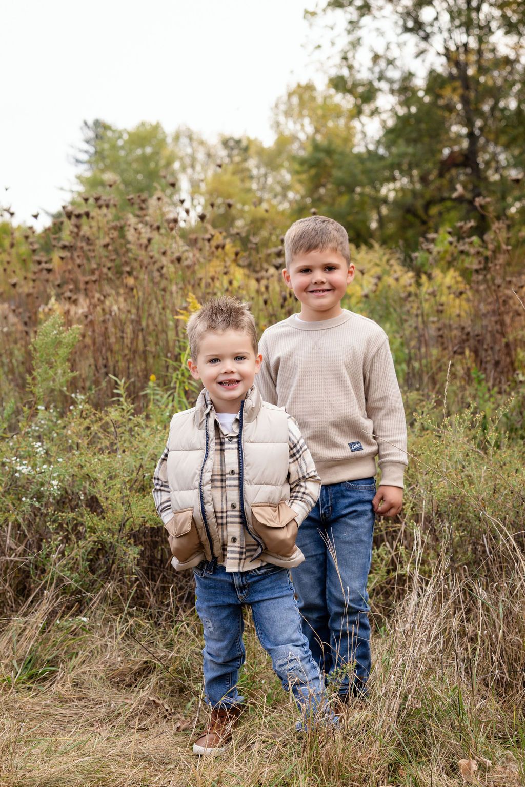 Two young boys smiling outdoors in fall, wearing jeans and casual tops, standing in a field with autumn foliage.