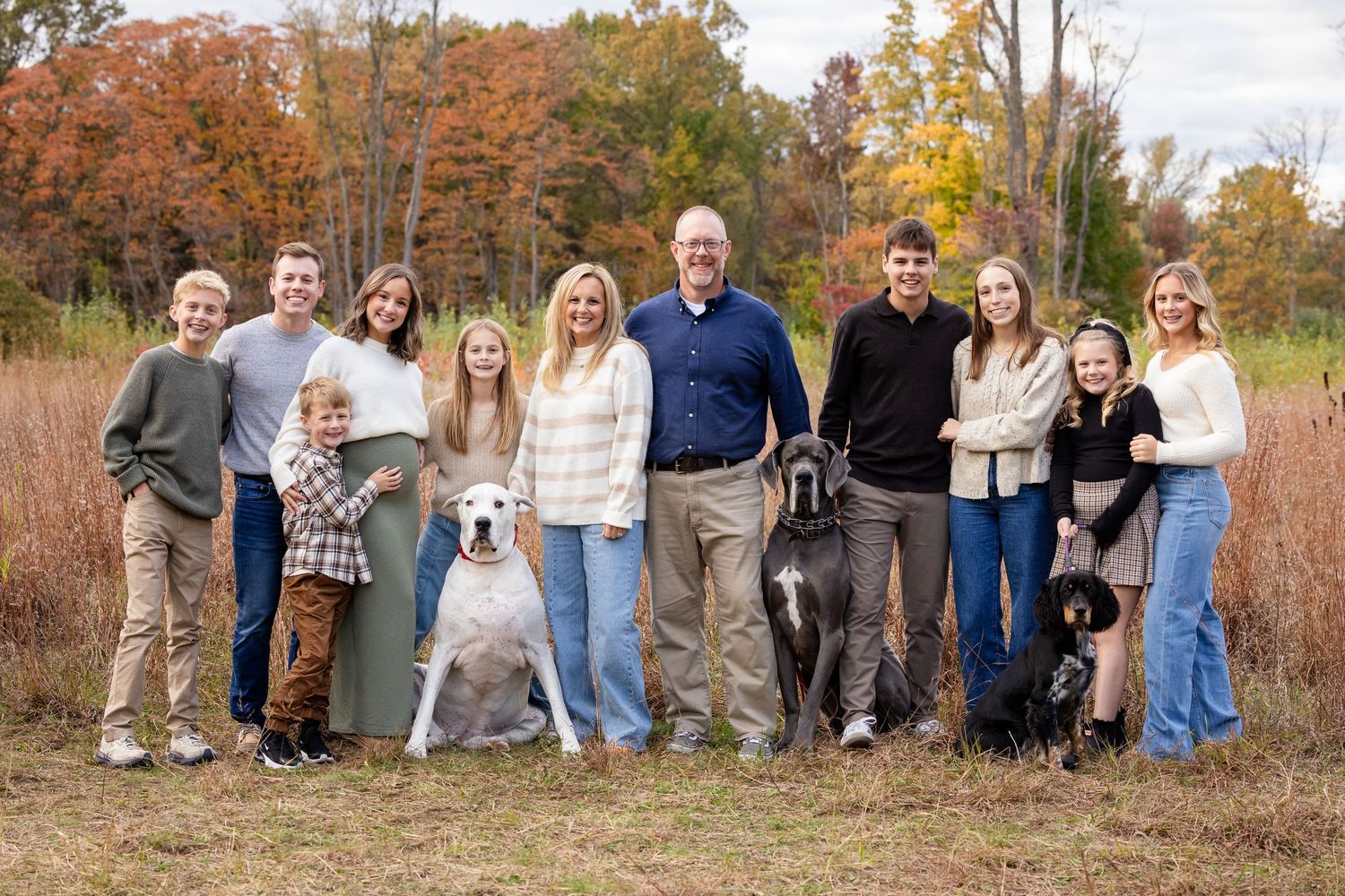 Large family group portrait with two dogs posing outdoors in autumn setting with colorful fall foliage background.