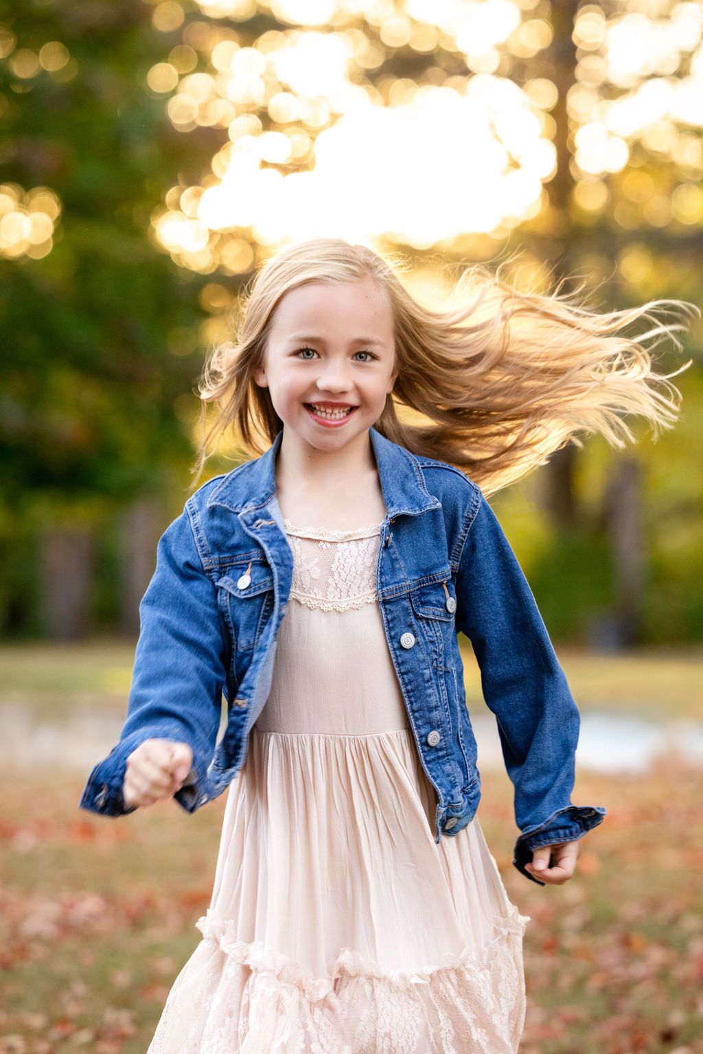 A joyful young girl with flowing blonde hair twirls outdoors in a denim jacket and pink dress with golden bokeh background.