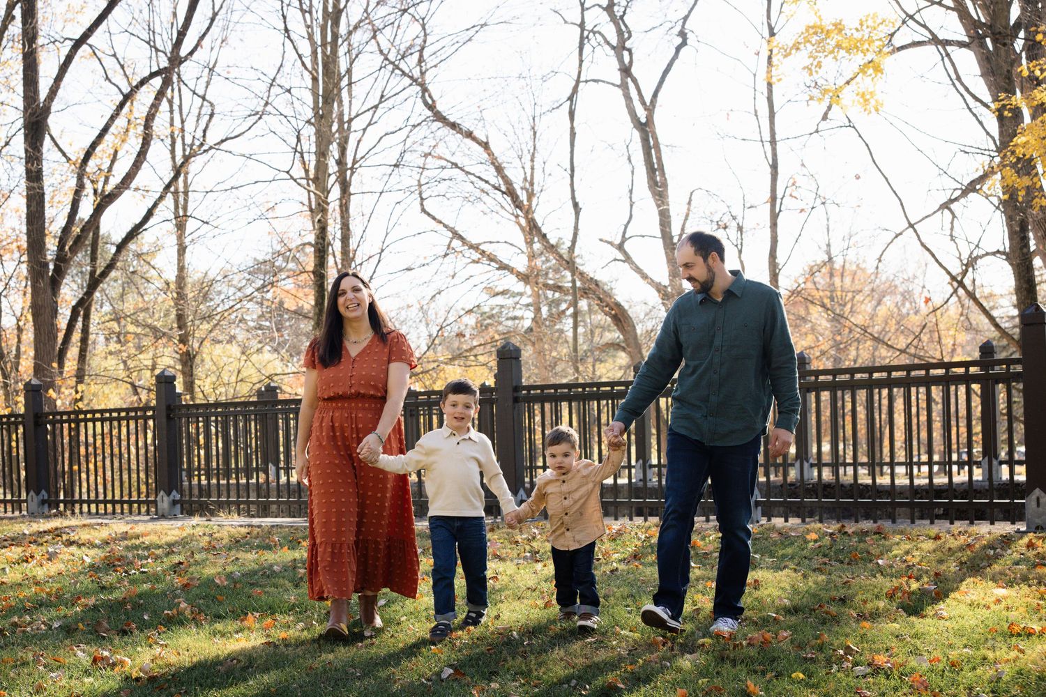 Family of four holding hands and walking in a park during autumn with colorful leaves and bare trees.