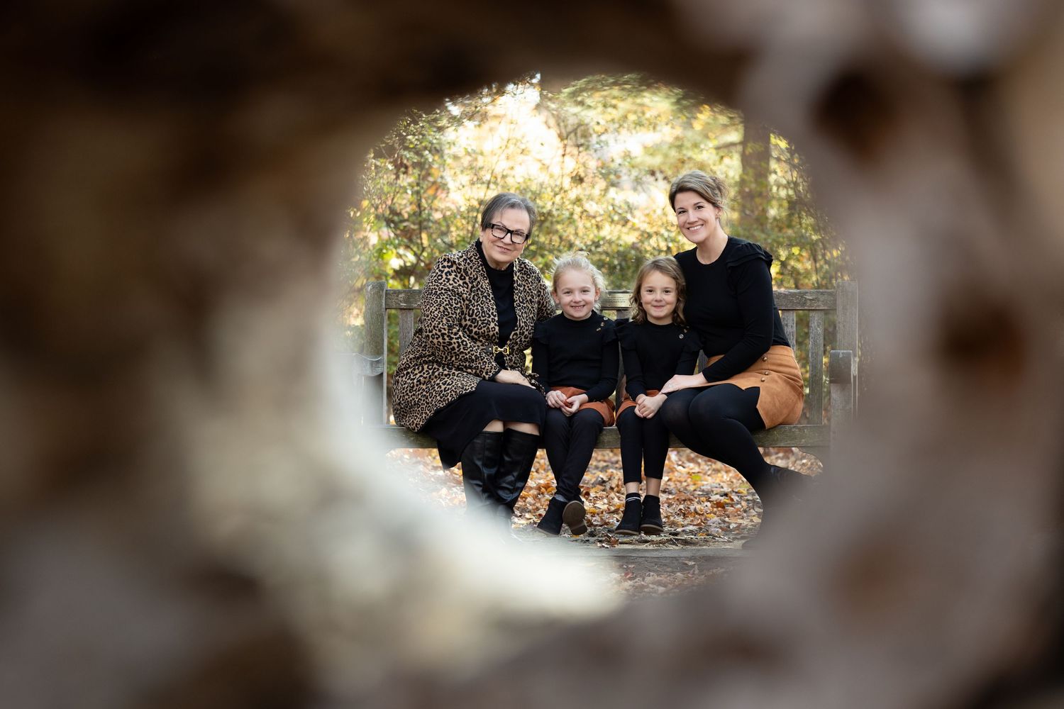 Family of four smiling outdoors in autumn, photographed through a hole in a tree trunk with fall foliage background.