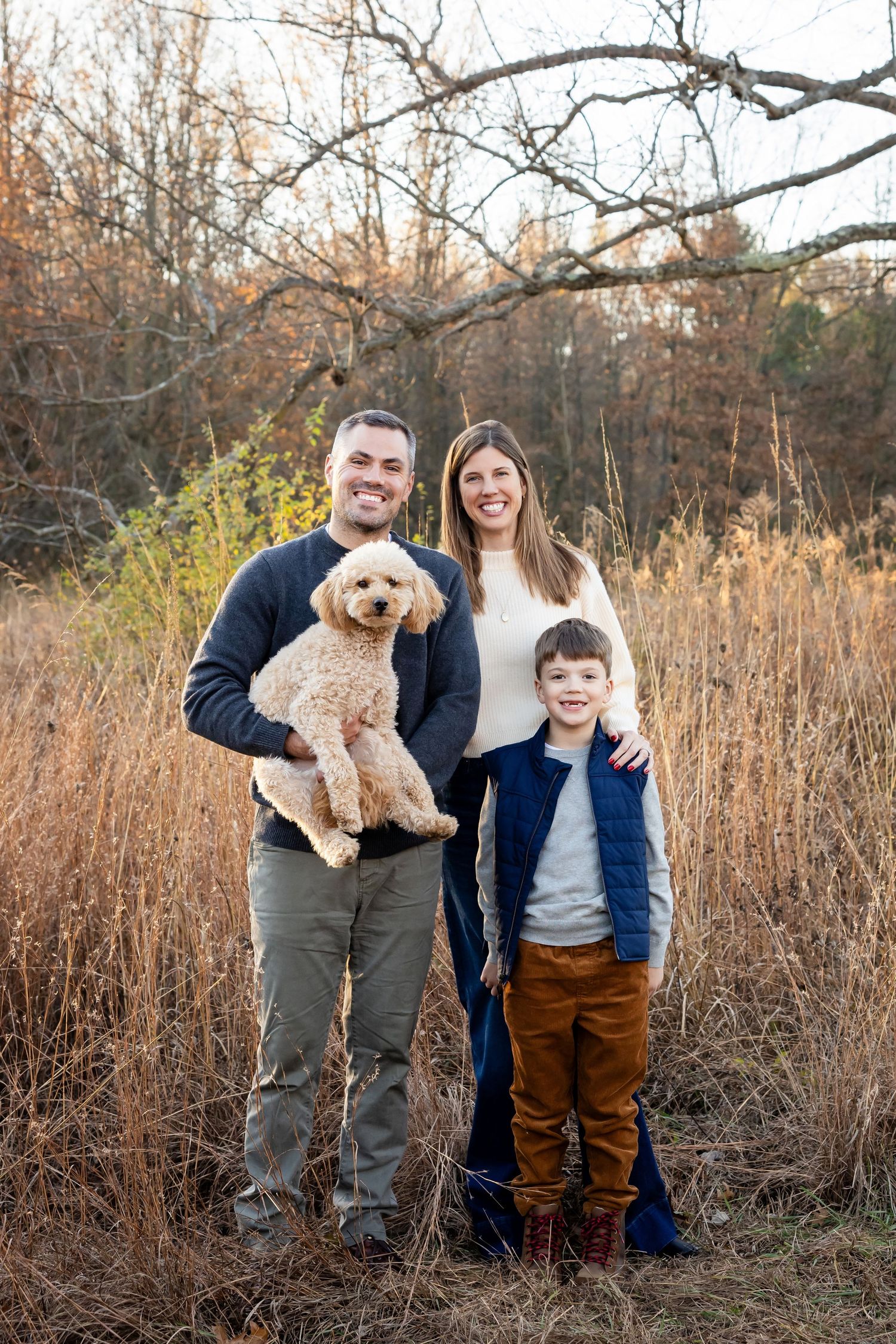 A smiling family of two adults and a child pose outdoors in a fall field, holding a fluffy golden doodle dog.