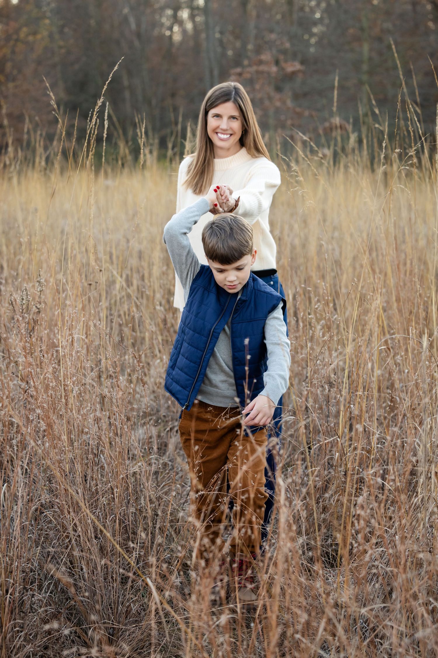 A smiling woman and young boy stand together in a golden autumn field with tall dry grass and trees.