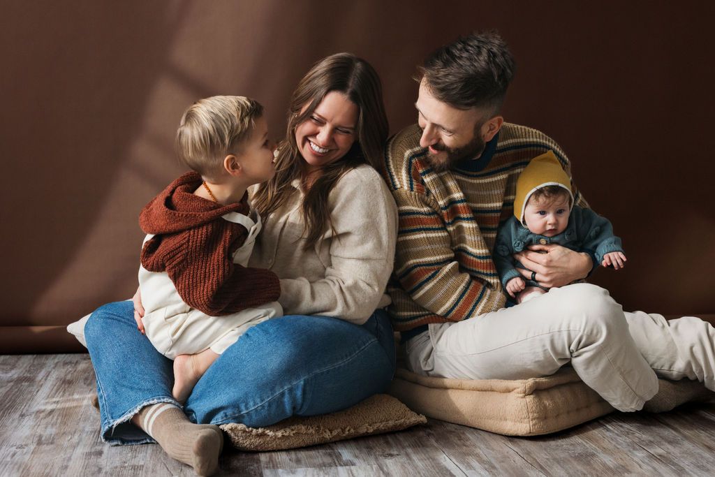 Happy family of four sitting on floor, parents smiling with toddler and newborn baby in warm autumn tones.