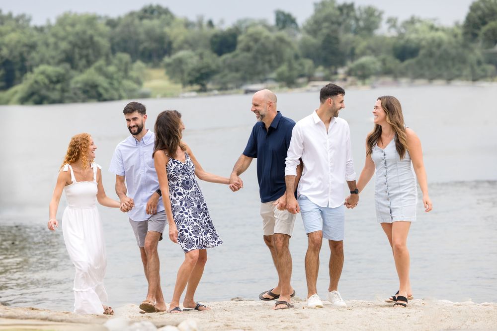 Six friends walking hand-in-hand along a sunny beach shoreline with lush green trees and a river in the background.