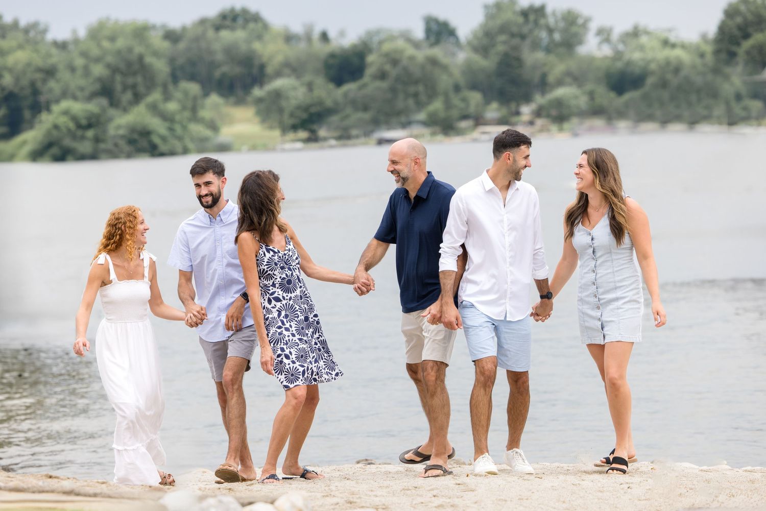 Six friends walking hand-in-hand along a sunny beach shoreline with lush green trees and a river in the background.