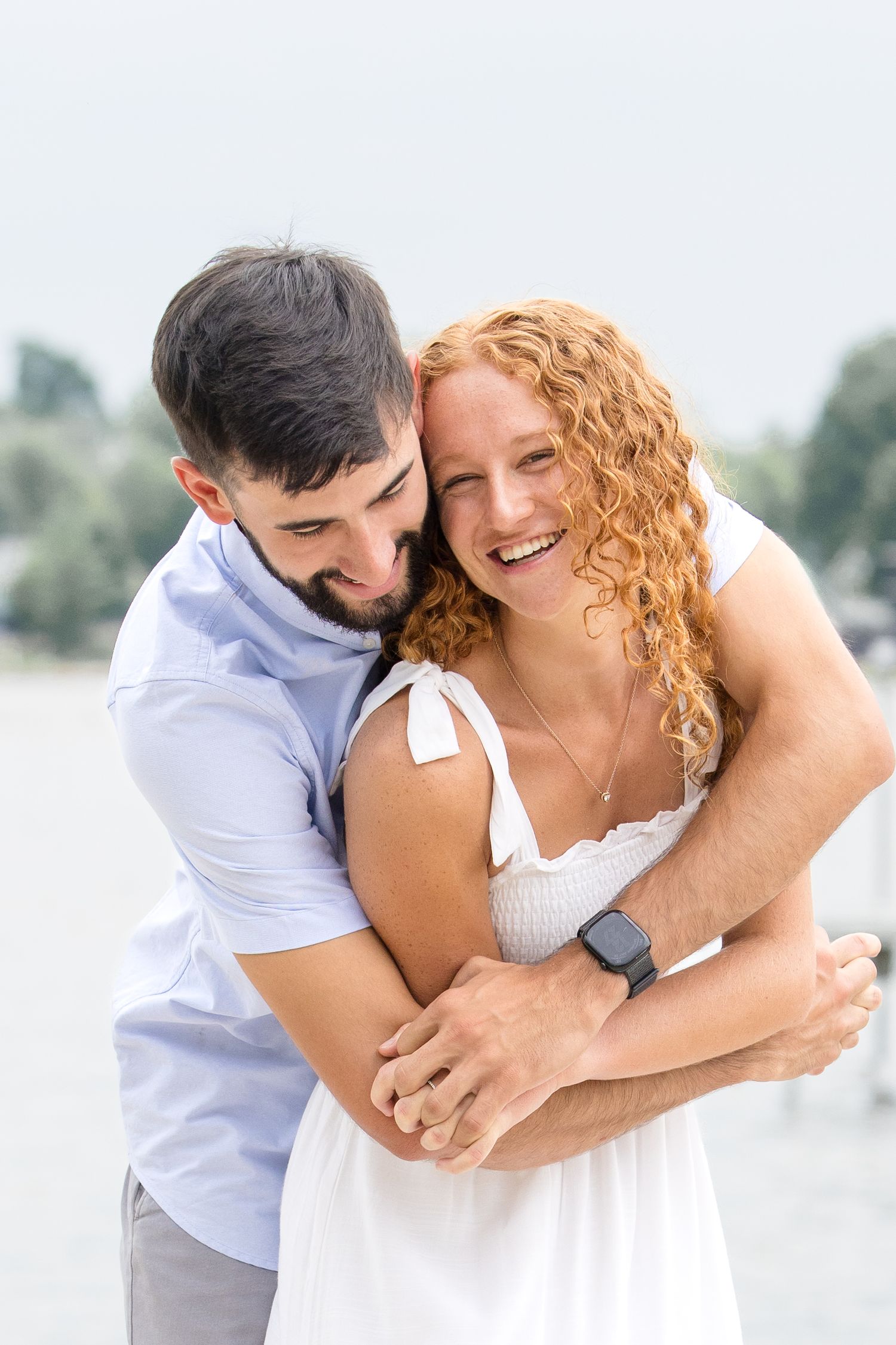 A happy couple embraces near a waterfront, the woman in a white dress laughing as the man hugs her from behind.