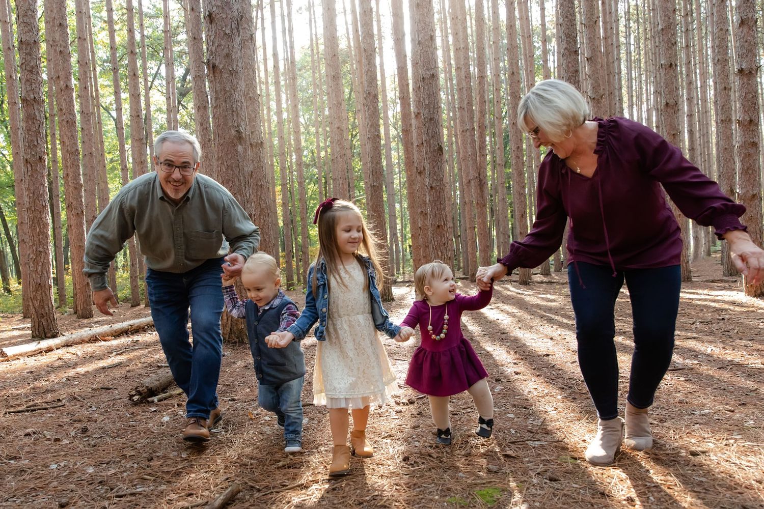 Grandparents walking with three young grandchildren through a sunlit pine forest, holding hands and smiling.