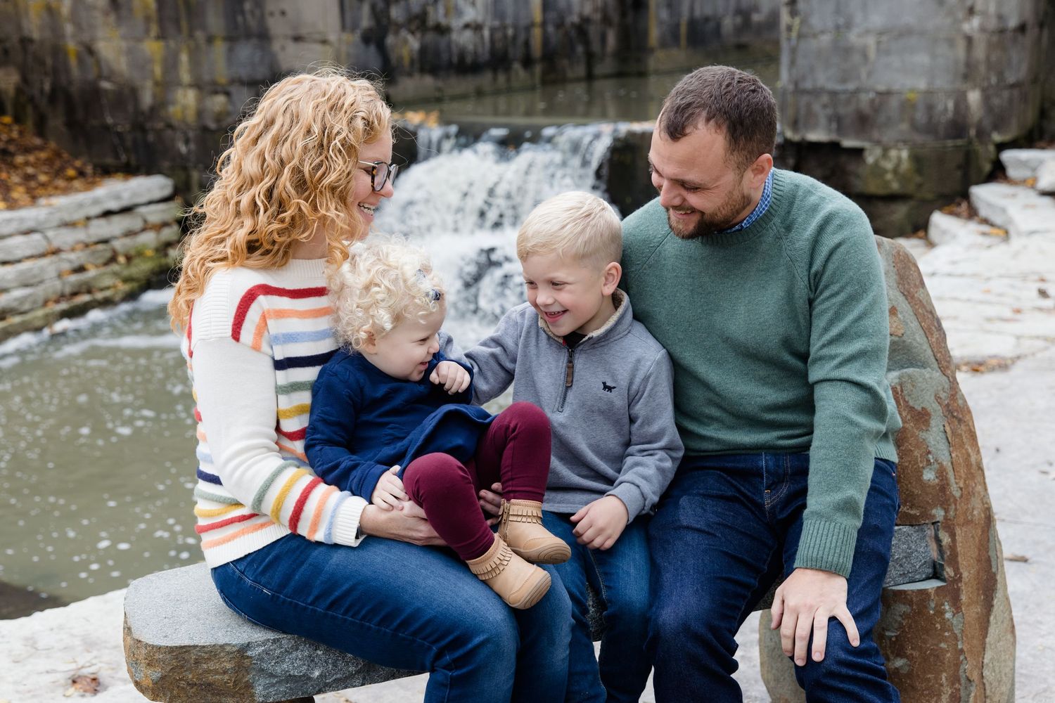 Family of four sitting on stone steps near a waterfall, smiling and enjoying a candid outdoor portrait session.
