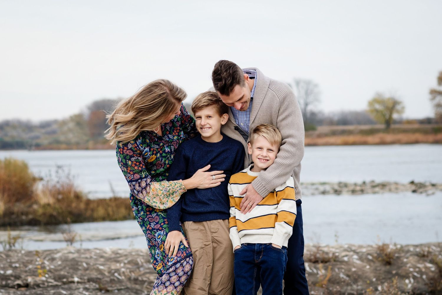 Family of four smiling and embracing outdoors near a lake during autumn, wearing casual clothes in a warm family portrait.