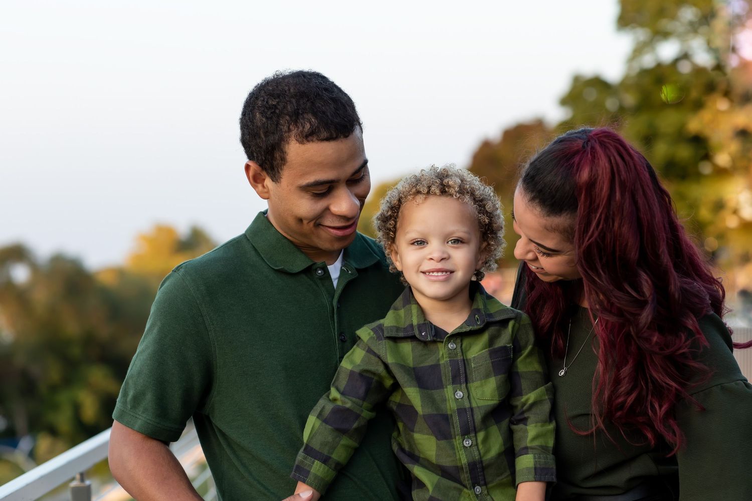 A happy mixed-race family with two parents and a young curly-haired toddler smiling together outdoors in autumn.