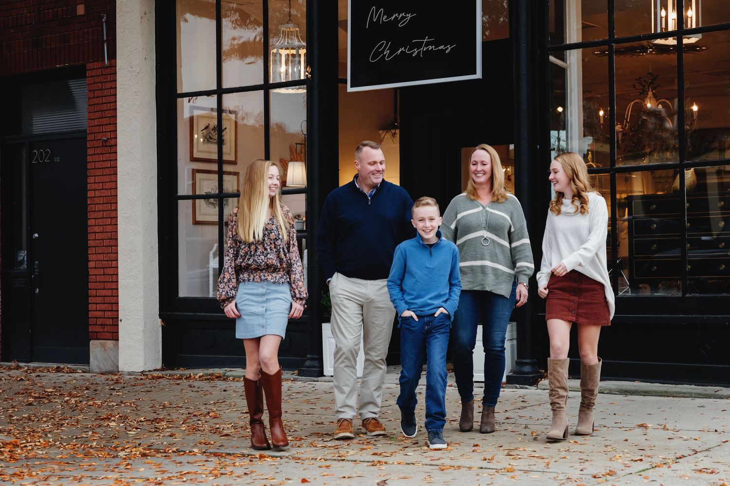 Family of five posing outdoors in fall attire in front of a store with a Merry Christmas sign.