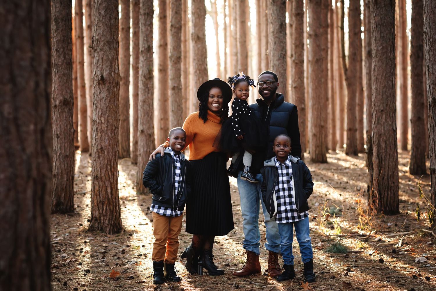Happy Black family posing together in a sunlit pine forest during fall, parents and three children smiling.