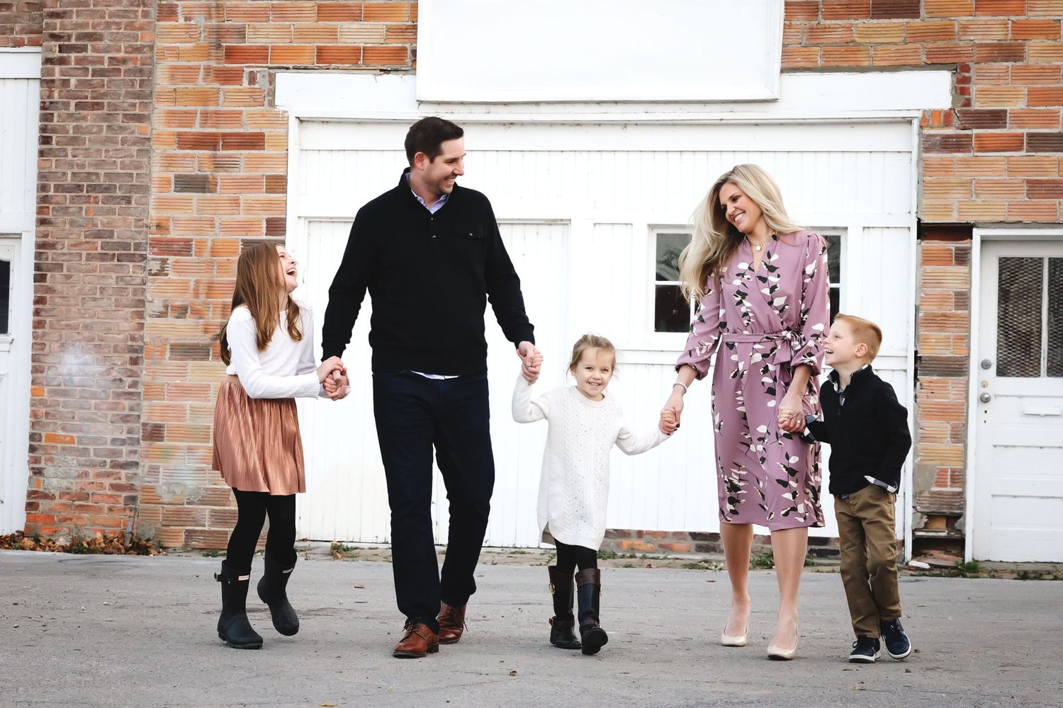 Happy family of five walking together outdoors, smiling and holding hands near a brick building.