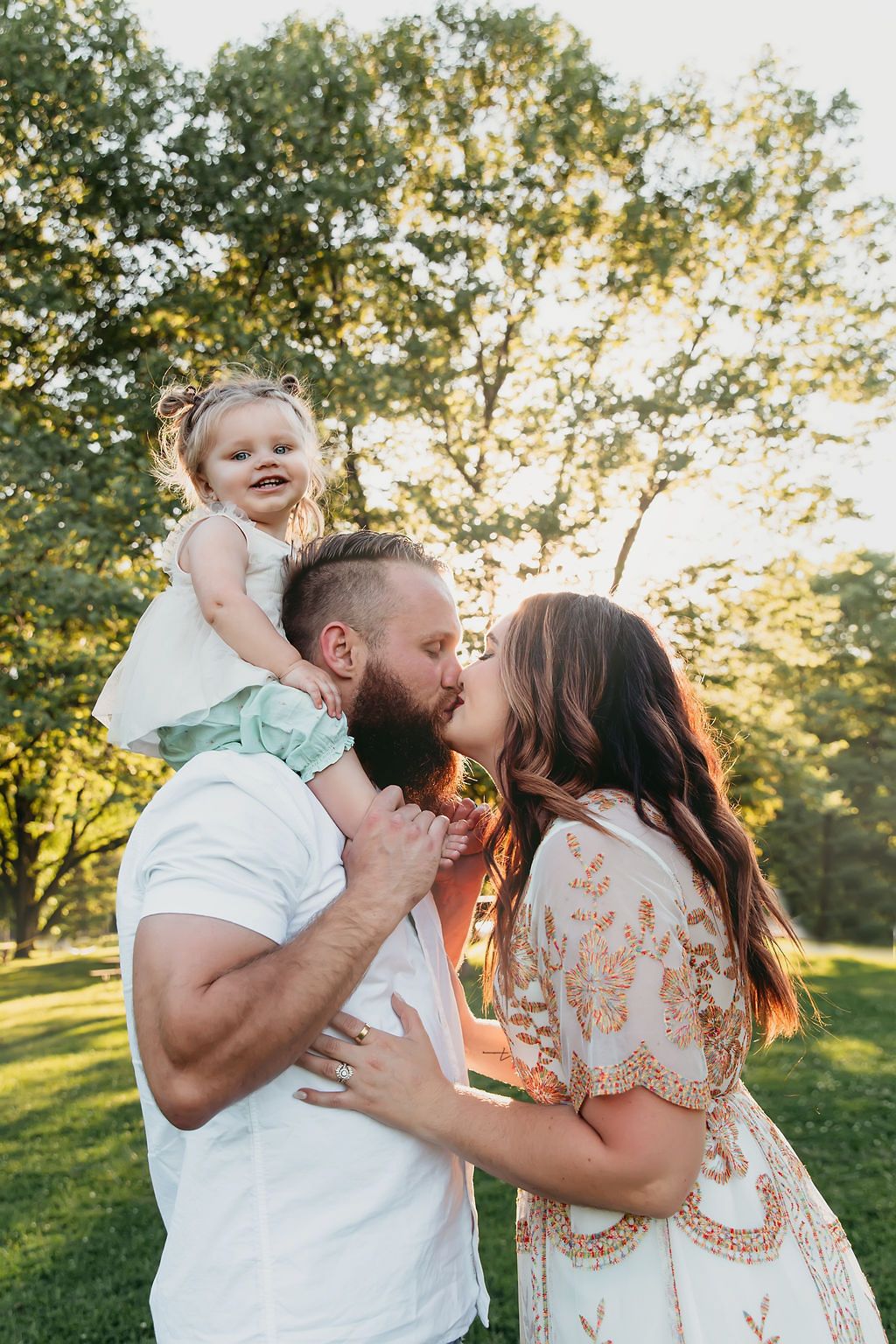 Couple kissing in sunny park while bearded father holds smiling toddler girl on his shoulders.
