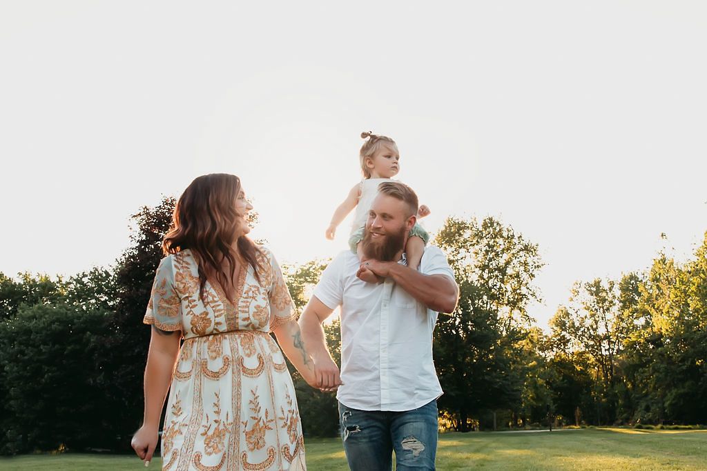 Happy couple walking in a sunny park, man carrying a toddler girl on his shoulders while holding hands.