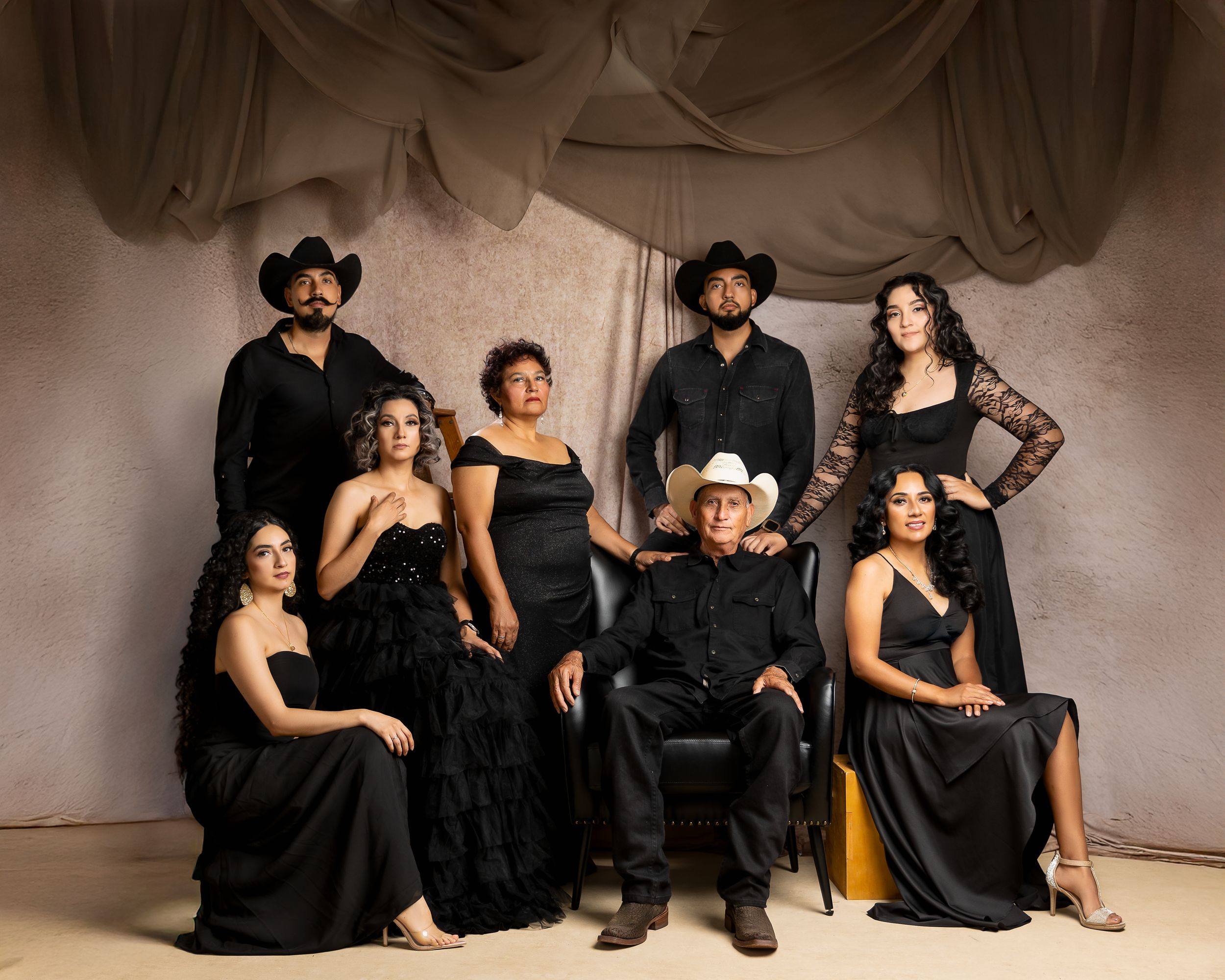 A group of seven people in elegant black outfits pose together in a studio with draped fabric backdrop.