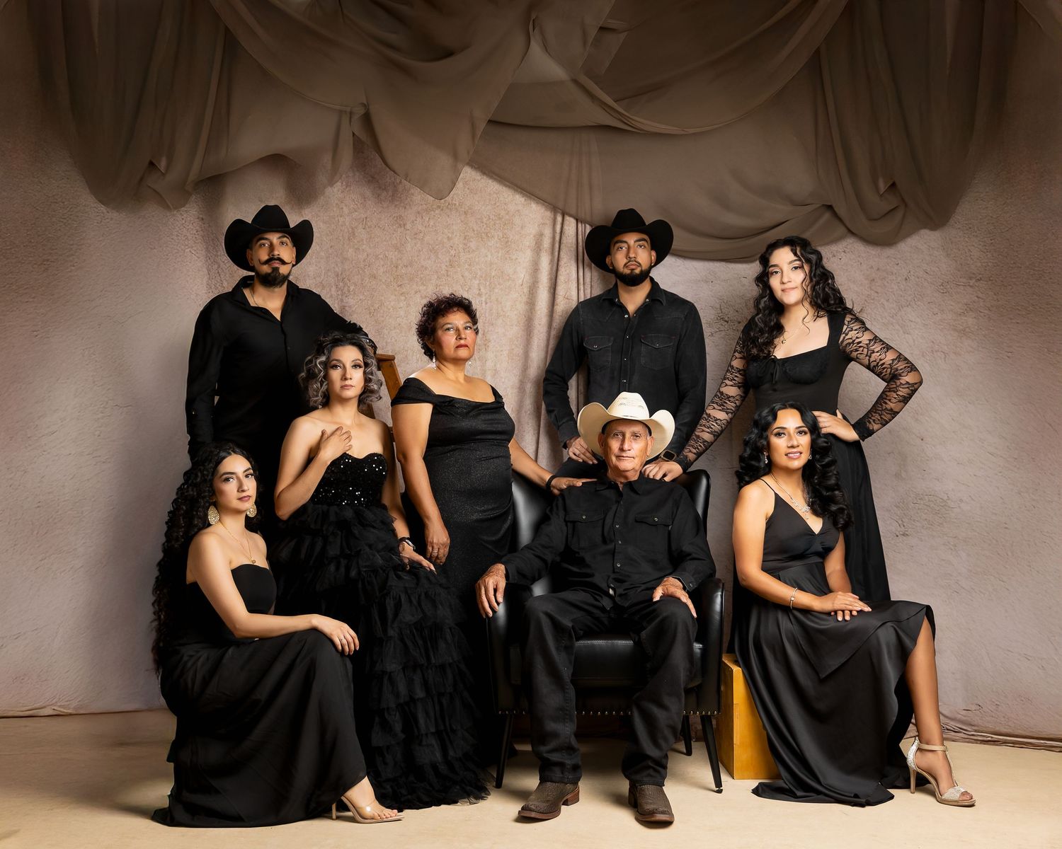 A group of seven people in elegant black outfits pose together in a studio with draped fabric backdrop.