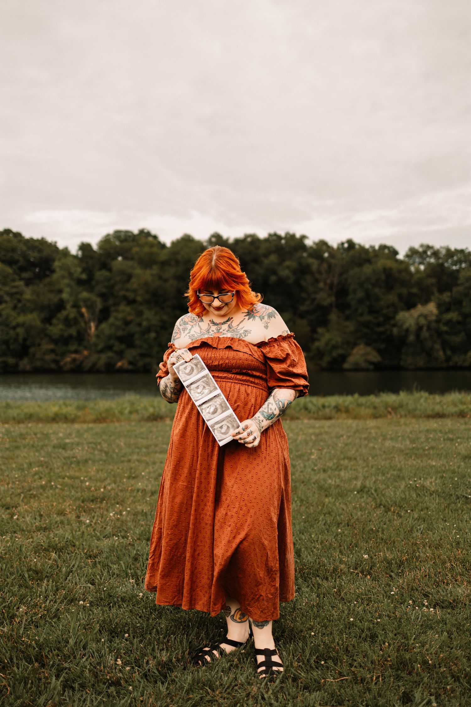 Pregnant woman in rust orange dress holding a sign outdoors near a lake, with red hair and tattoos.