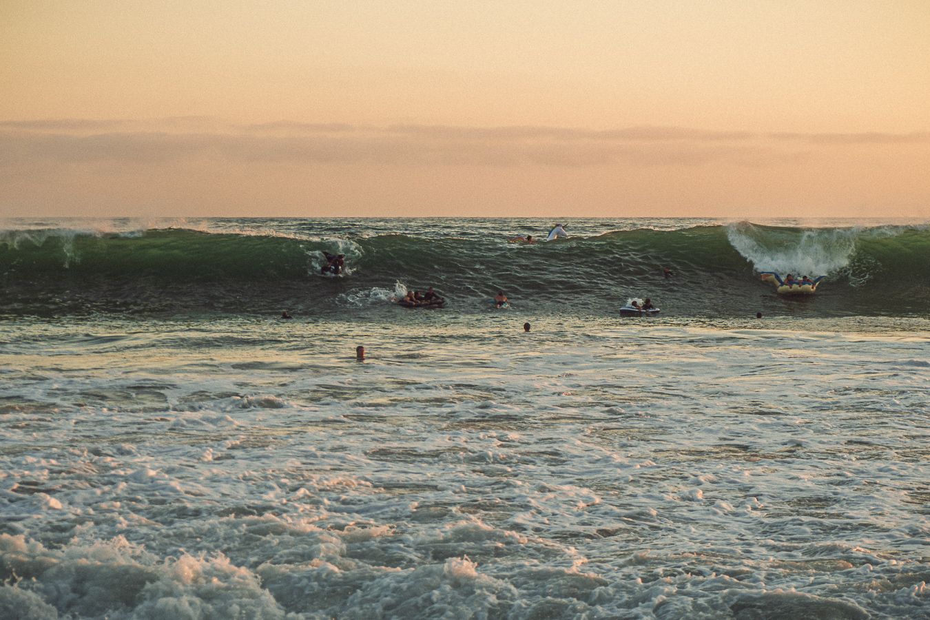 Surfers riding large ocean waves at sunset with golden sky and white foam crashing on the shore.
