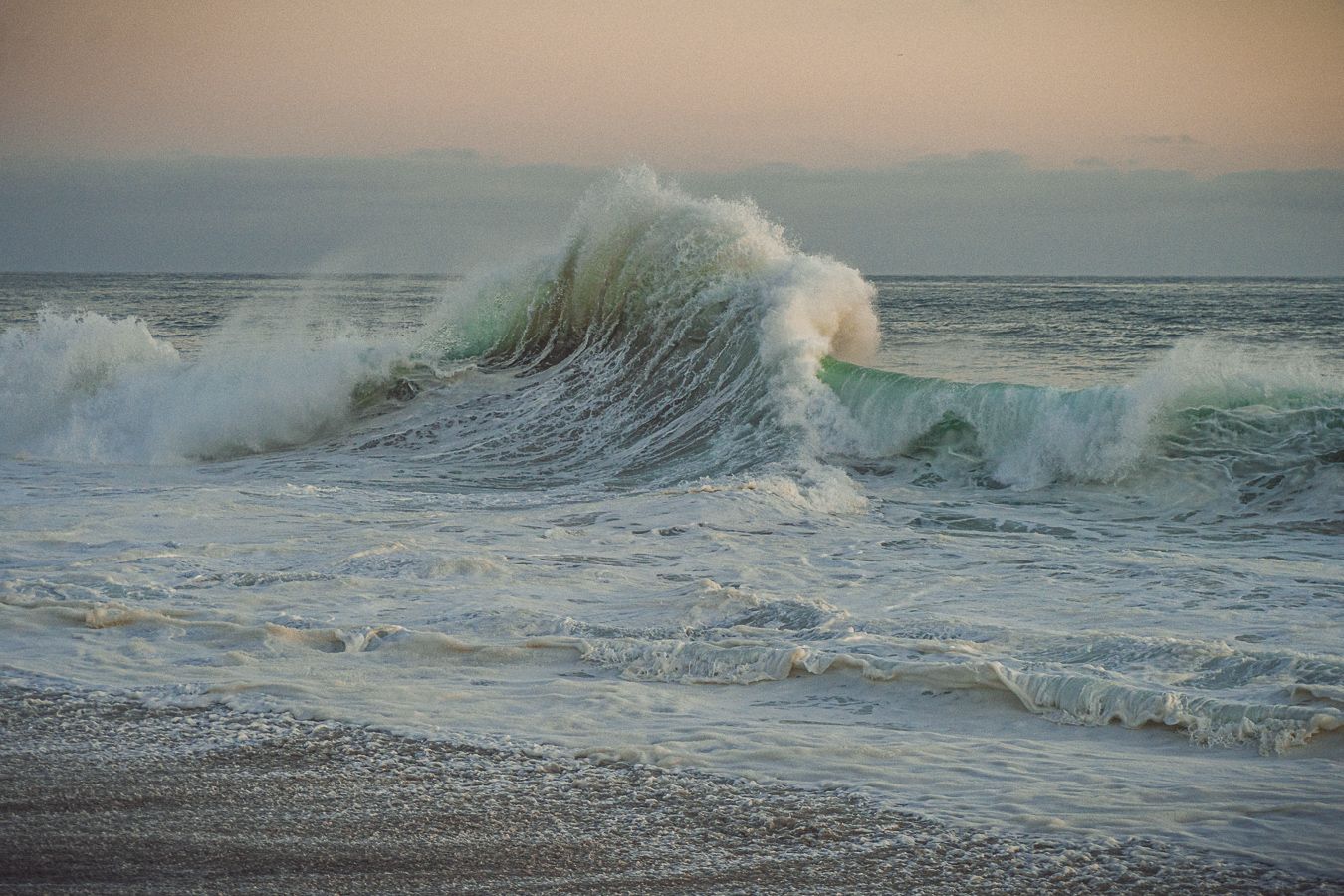 Large powerful ocean wave cresting and breaking on a sandy beach at dusk.