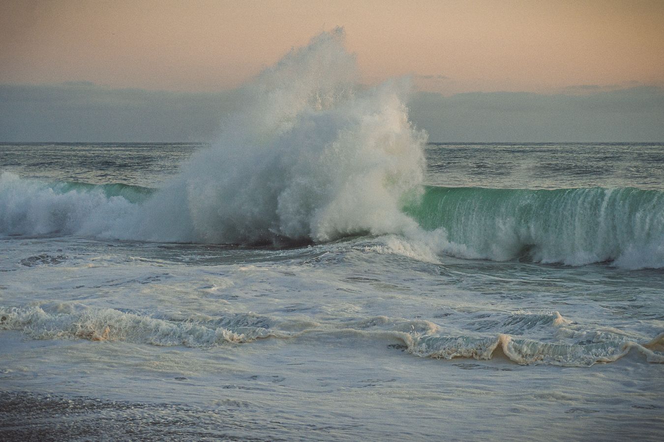 Powerful ocean wave crashing dramatically against the sea surface during a colorful sunset.
