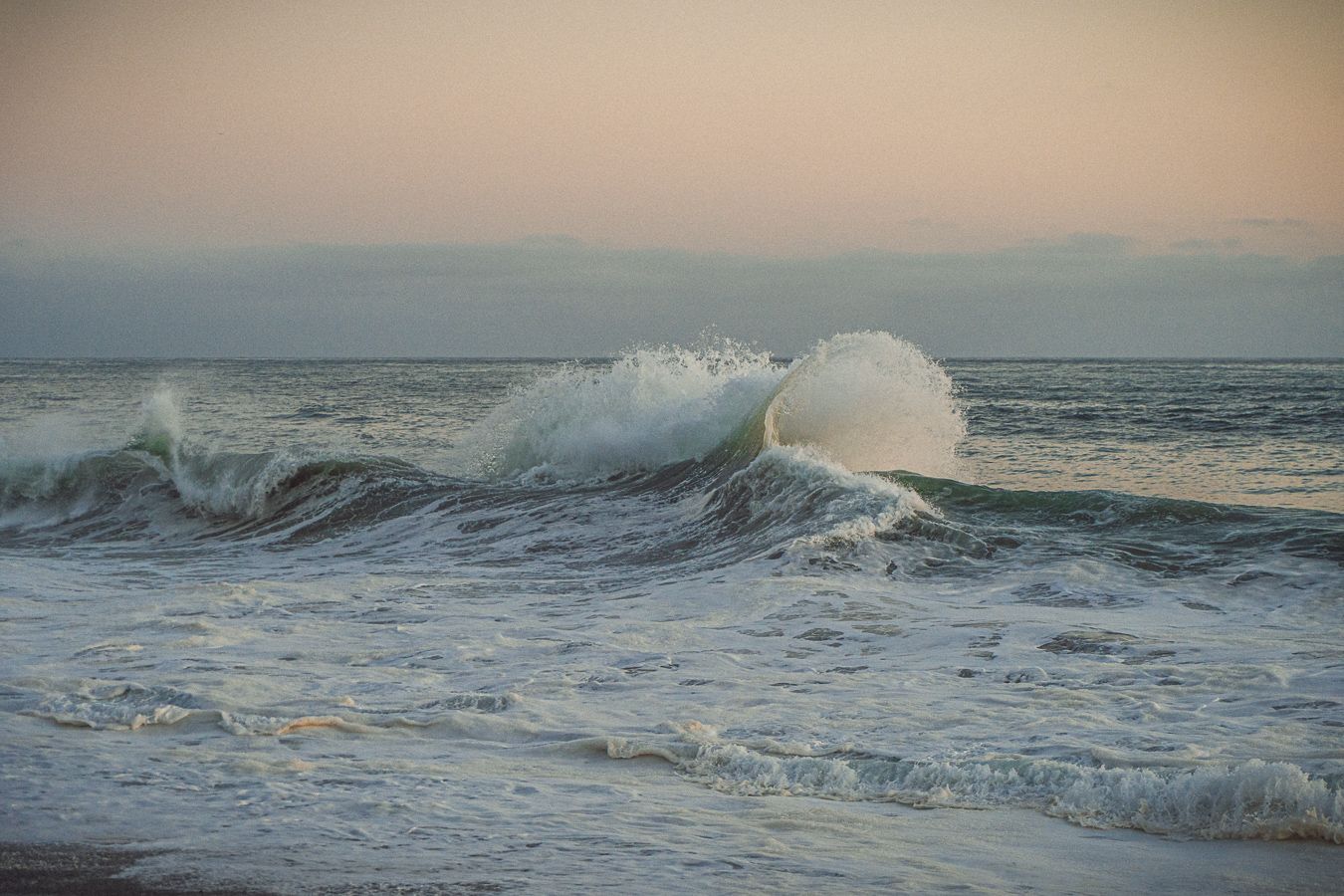 Ocean waves crashing at sunset with seafoam and mist rising above the water on a calm evening beach.