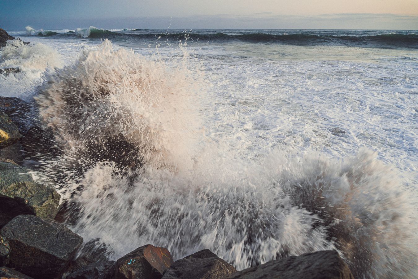 Powerful ocean wave crashing against rocky shoreline, sending white sea foam and water spray into the air.