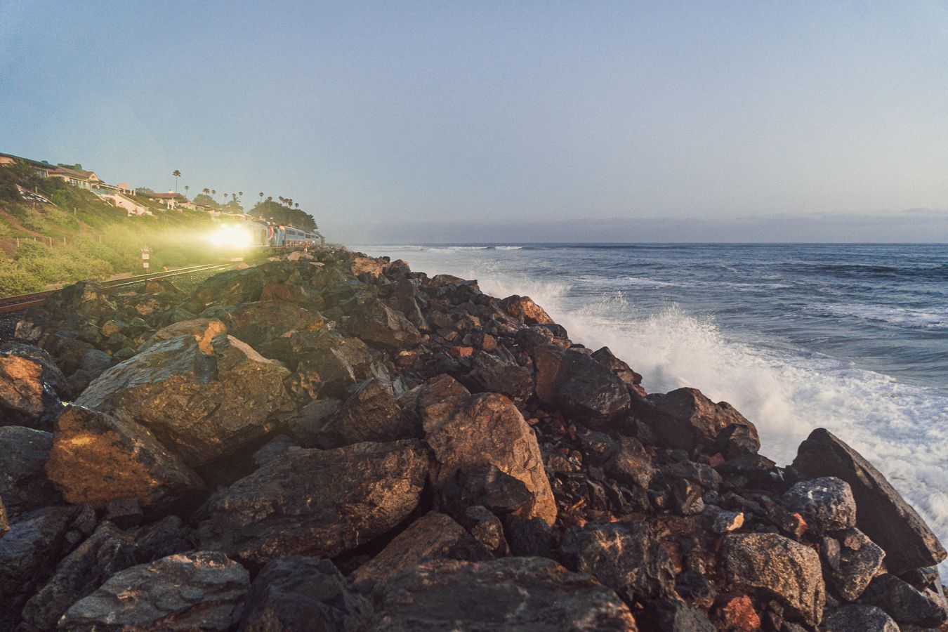 Rocky coastal jetty with waves crashing at dusk, glowing light visible on a grassy cliff in background.