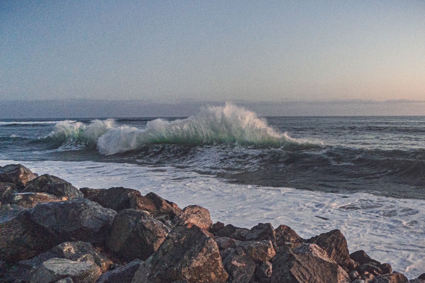 A large green wave cresting and breaking near a rocky shore during a calm coastal sunset.