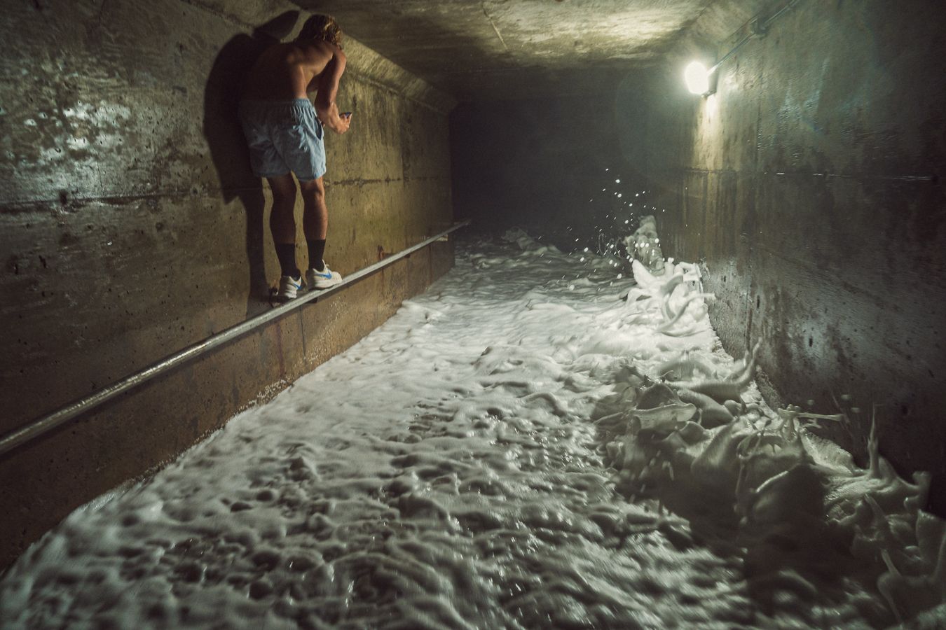 Two people stand on a ledge in a flooded underground tunnel with rushing white water below and dim lighting.