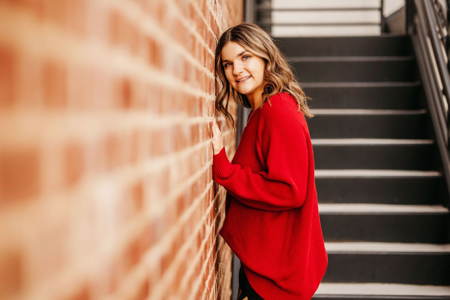 Young woman in red sweater smiling while leaning against a brick wall near outdoor stairs.