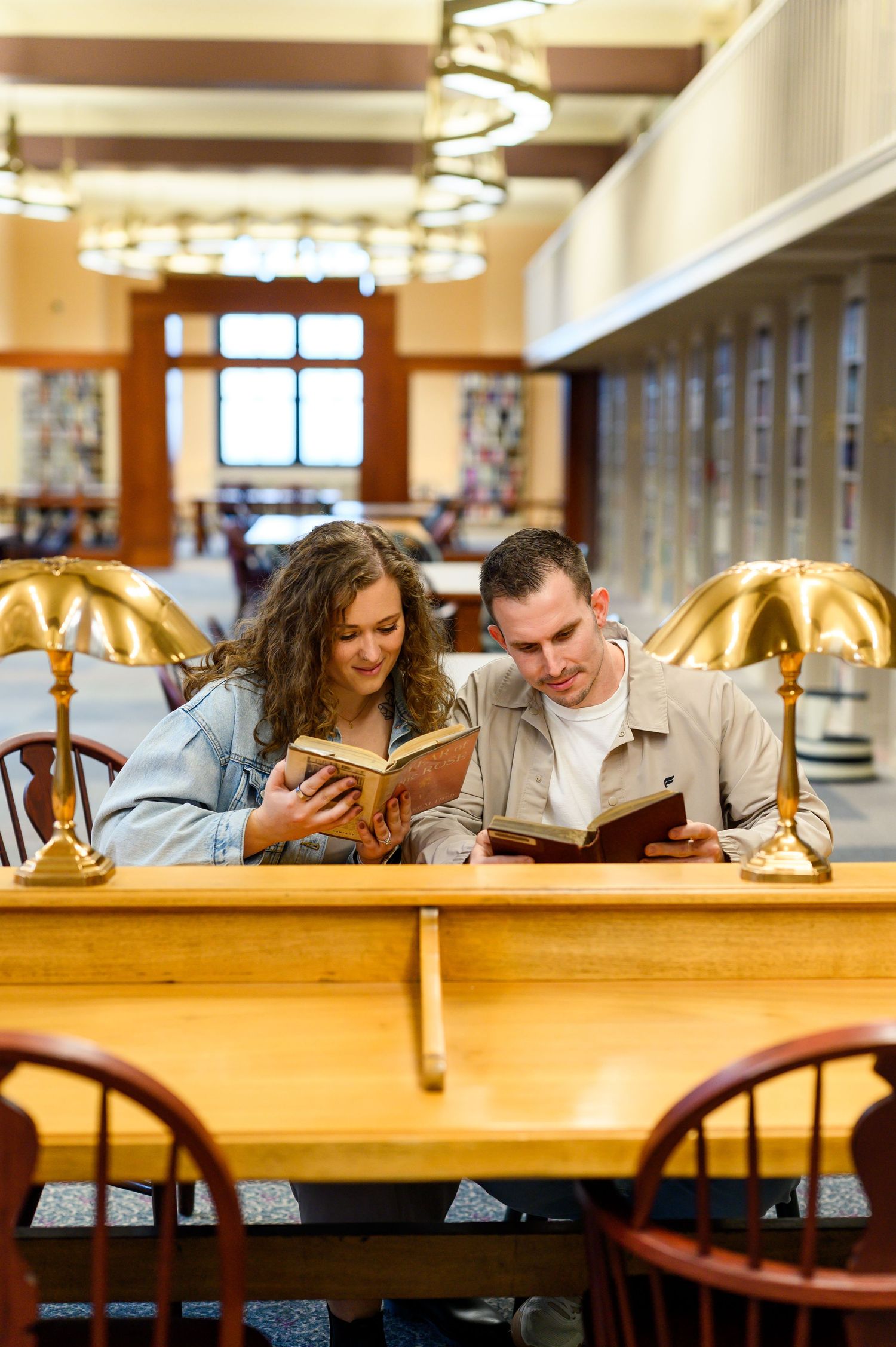 Two students reading books together at a wooden library table with brass lamps in an elegant academic library.