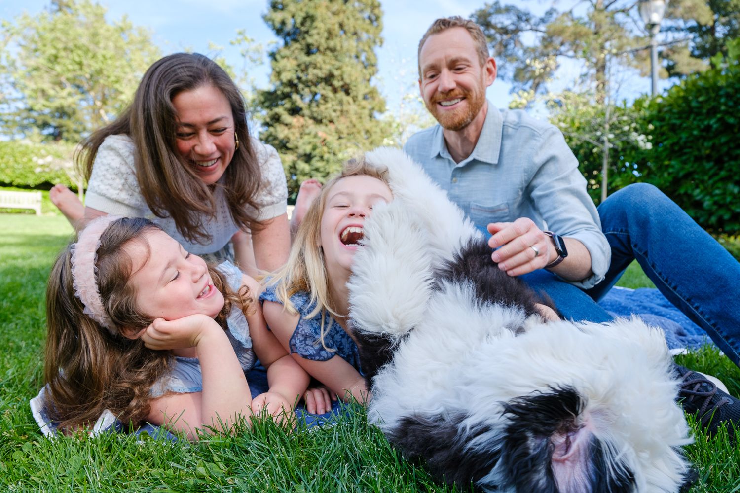 Happy family with two girls and a fluffy dog laughing together on green grass in a sunny park.