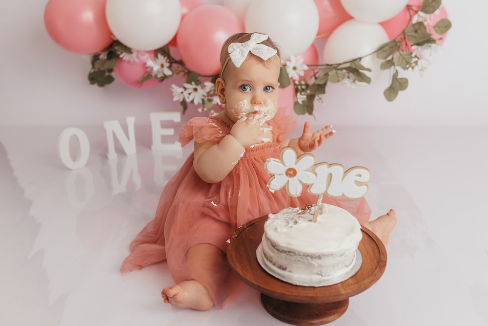 Baby girl in pink dress enjoys first birthday cake smash with ONE decorations and pink balloons backdrop.